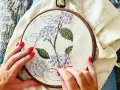 Manos con uñas rojas bordando una flor de hortensia en un bastidor. Se ve lo que se necesita para bordar: tela, hilo y aguja.