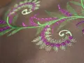 Detalles de bordado floral en tonos verdes y morados sobre tela marrón. Ideal para descargar letras para bordar a maquina y crear diseños únicos.