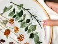 Manos creando bellas ideas para bordar: hojas verdes y flores naranjas en tela, con aguja e hilo listos para continuar.