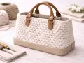 Bolso tejido a mano con asas de cuero. La tela para bolsos es de color crema, con base de cuero beige. A su lado, ovillos de lana y herramientas de costura.