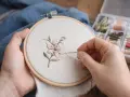 Manos delicadas bordando una flor con hilo rosa y verde sobre tela clara, usando un bastidor. A un lado, una caja con carretes de hilo.