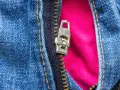 Detalle de cremallera metálica en jeans azules, con un forro rosa vibrante que se asoma. La cremallera salida de un lado revela el contraste de texturas.