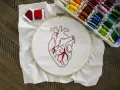 Un corazón bordado en tela blanca, listo para aprender a bordar. Hilos rojos y una caja de hilos de colores completan la escena.