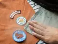 Tres parches decorativos sobre tela marrón. Una mano presiona tela verde con una plancha, para saber si un parche es termoadhesivo.