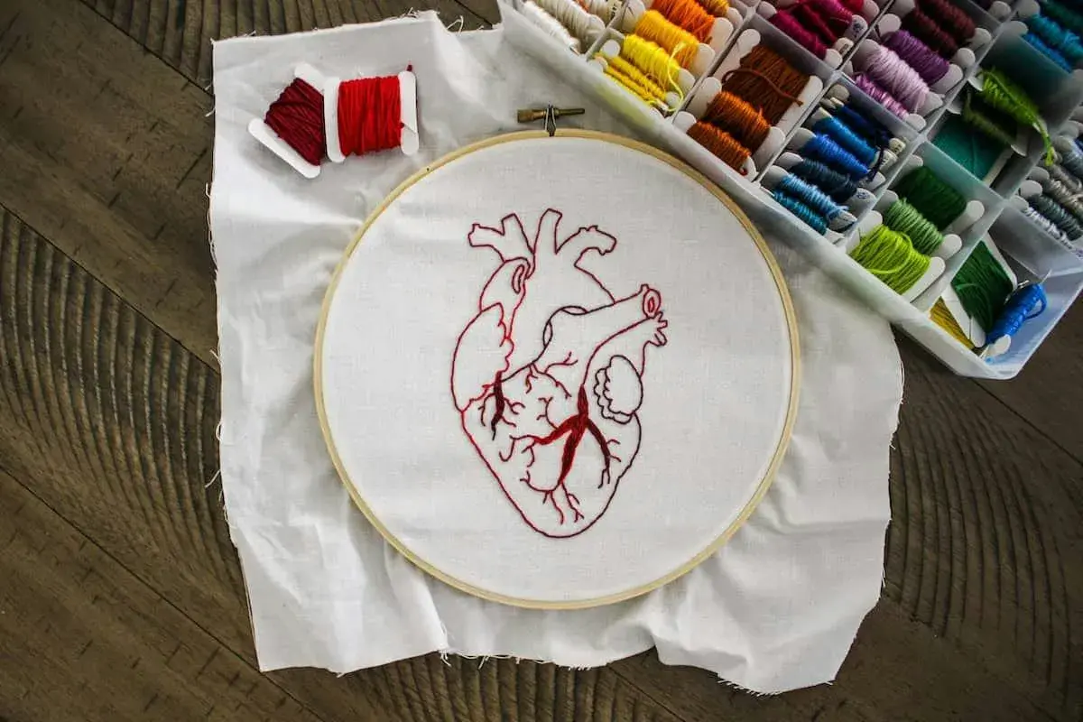 Un corazón bordado en tela blanca, listo para aprender a bordar. Hilos rojos y una caja de hilos de colores completan la escena.