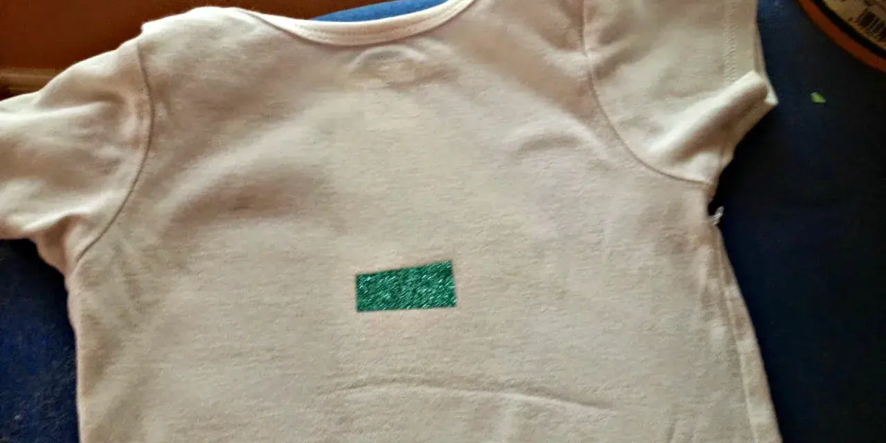 Camiseta blanca con un pequeño rectángulo verde brillante en el centro. Aprende como quitar pegatinas termoadhesivas de la ropa.