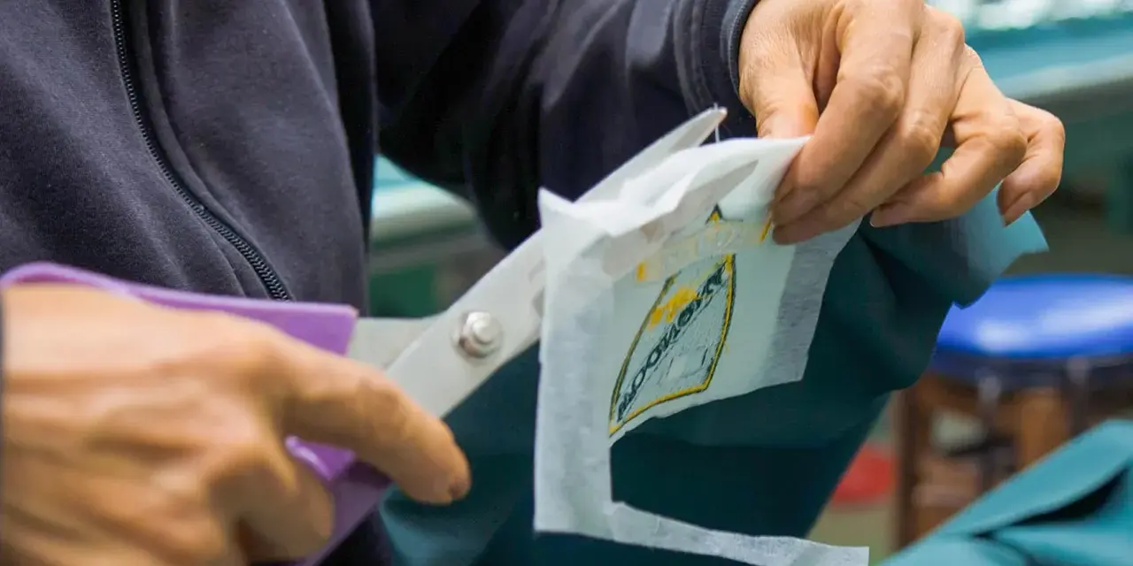 Manos cortan con tijeras un parche bordado sobre tela estabilizadora, preparándolo para ser cosido.