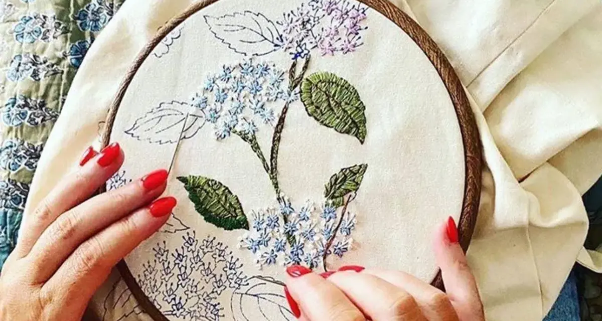 Manos con uñas rojas bordando una flor de hortensia en un bastidor. Se ve lo que se necesita para bordar: tela, hilo y aguja.