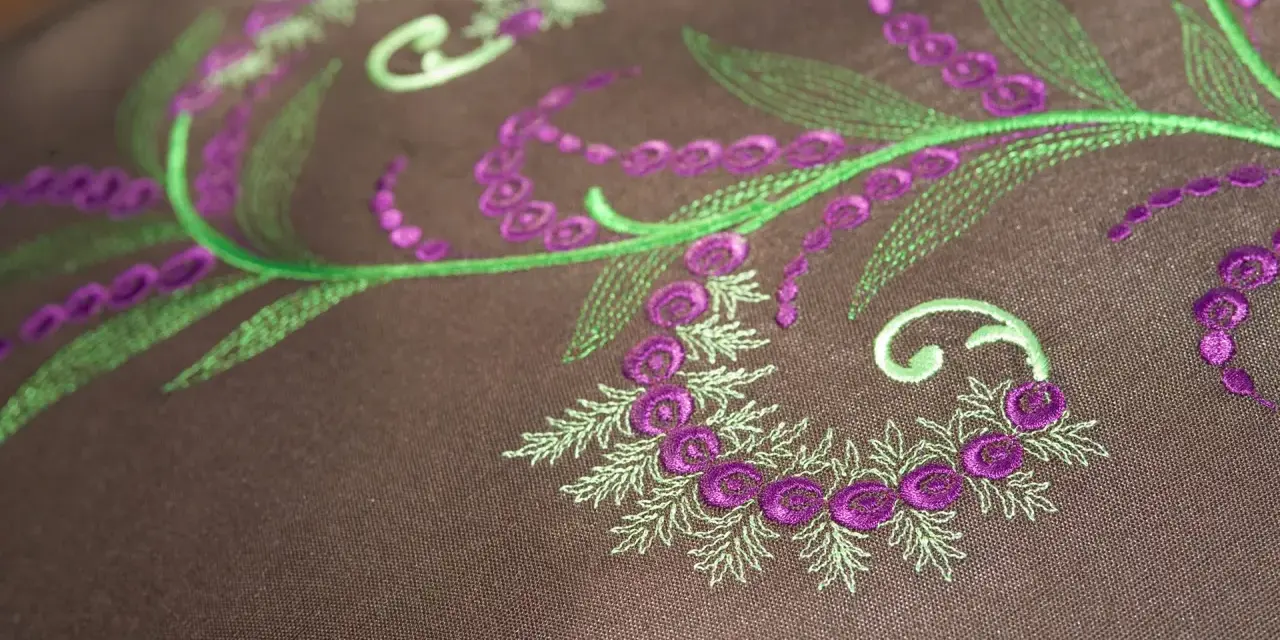 Detalles de bordado floral en tonos verdes y morados sobre tela marrón. Ideal para descargar letras para bordar a maquina y crear diseños únicos.