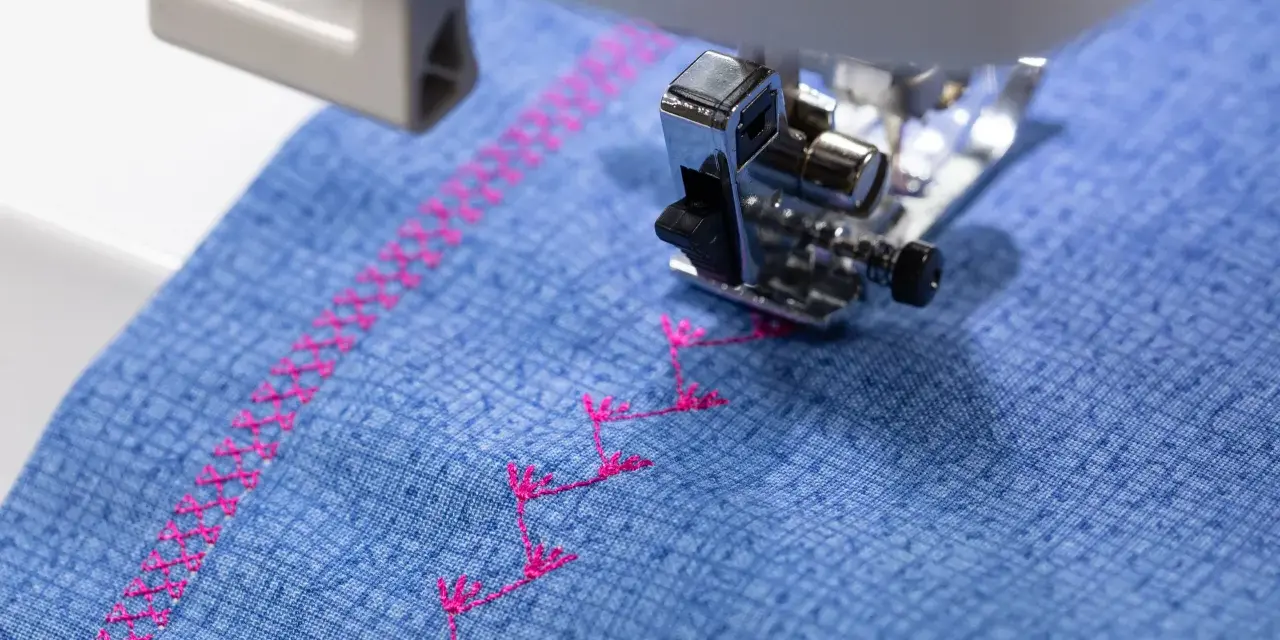 Máquina de coser bordando un patrón decorativo en **tejidos de algodón** azul con hilo rosa.