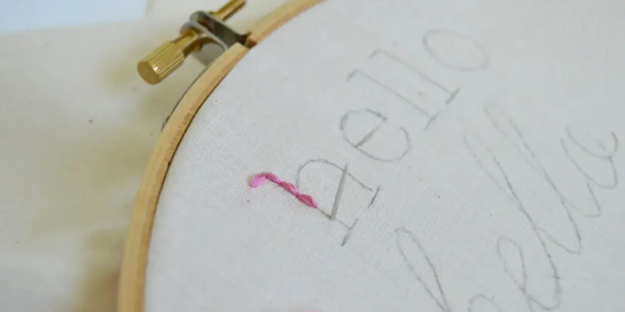 Bastidor con tela y un hilo rosa comenzando a bordar letras.