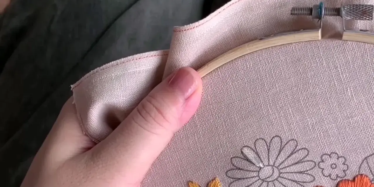 Mano sujetando un bastidor de bordar con lino para bordar. Se aprecian flores a medio bordar en tonos grises, naranjas y amarillos.