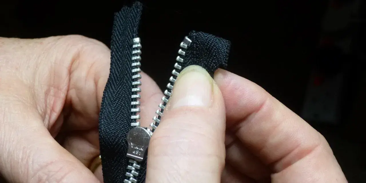 Manos sujetan una cremallera negra, alineando los dientes para coser cremallera a mano.