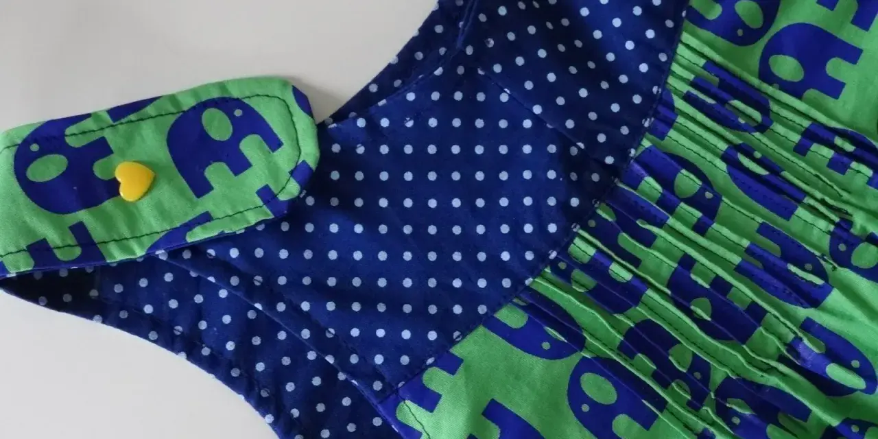 Detalle de un vestido infantil con estampado de elefantes azules sobre fondo verde. El cuello tiene un detalle de lunares azules y un botón amarillo en forma de corazón. Un adorable **patrón ranita bebé**.