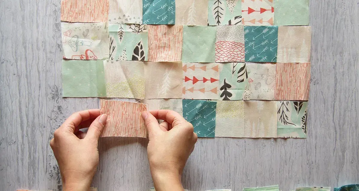 Manos uniendo retazos de tela con diseños de árboles y flechas, creando un patrón de patchwork.