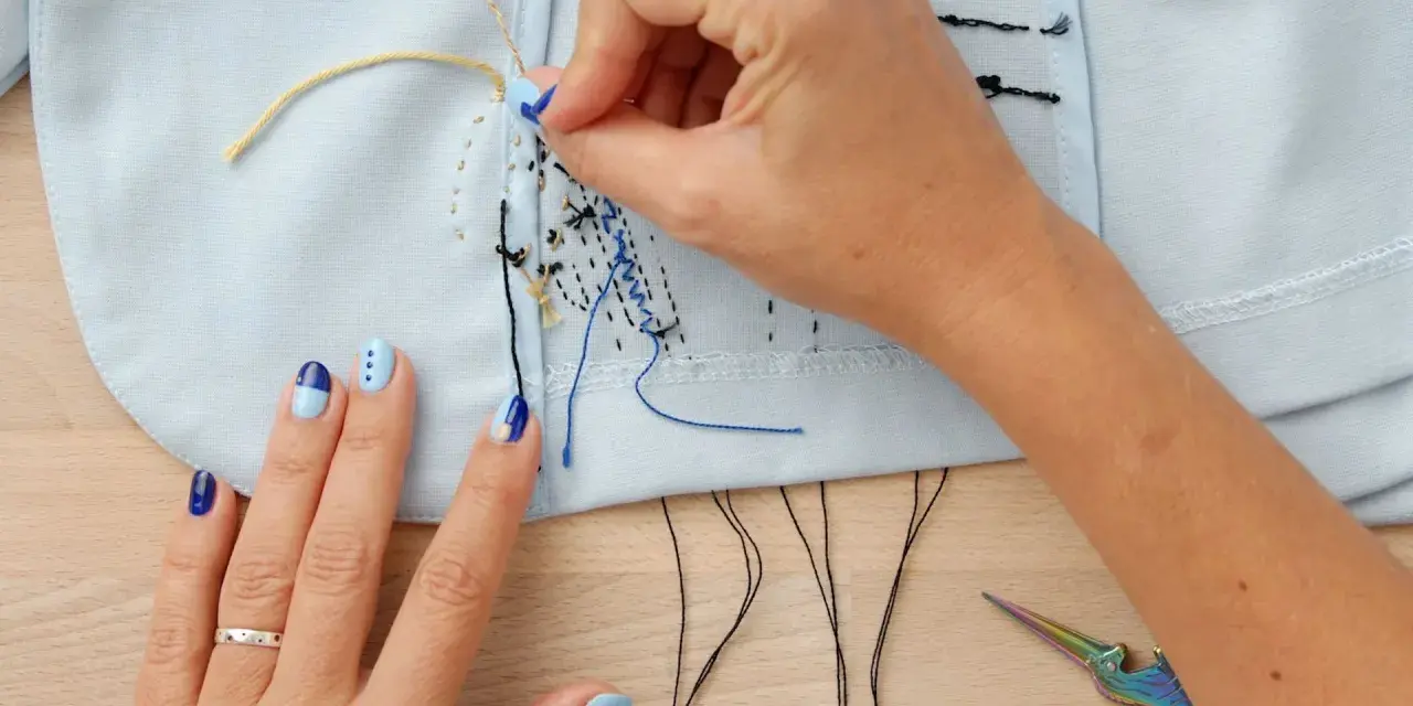 Manos con uñas decoradas bordando con hilo de coser sobre tela azul claro. Tijeras de colores y hilos negros a la vista.