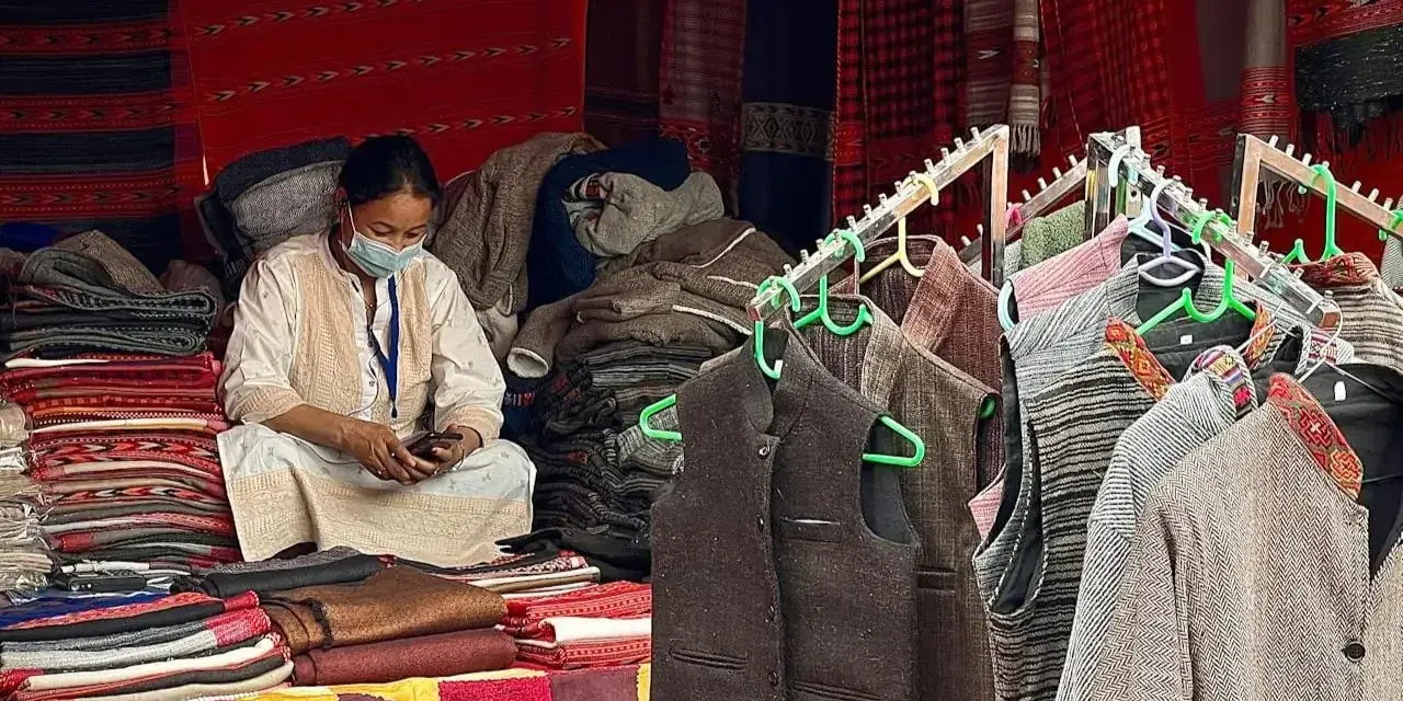Mujer enmascarada sentada entre pilas de ropa de lana cachemira. Chalecos y chaquetas de lana cuelgan en perchas.