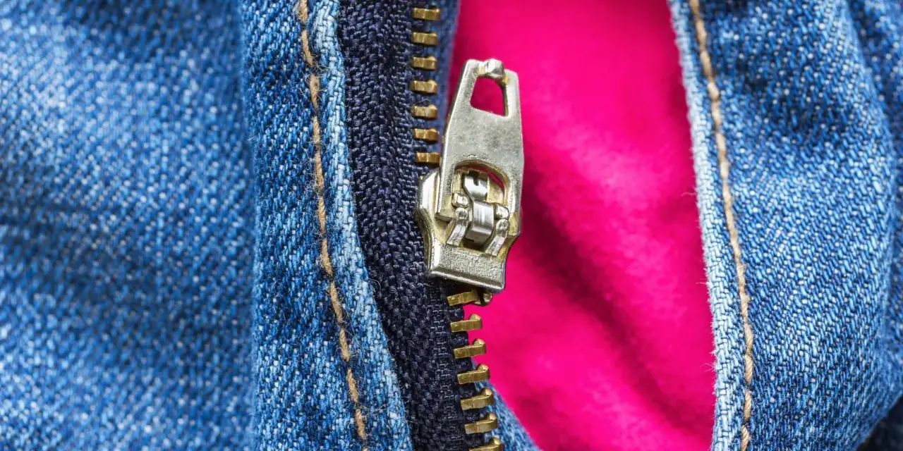 Detalle de cremallera metálica en jeans azules, con un forro rosa vibrante que se asoma. La cremallera salida de un lado revela el contraste de texturas.