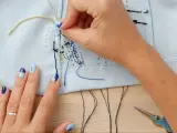 Manos con uñas decoradas bordando con hilo de coser sobre tela azul claro. Tijeras de colores y hilos negros a la vista.