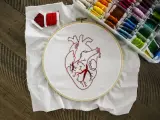 Un corazón bordado en tela blanca, listo para aprender a bordar. Hilos rojos y una caja de hilos de colores completan la escena.