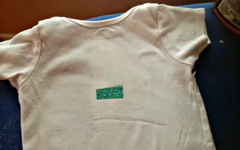 Camiseta blanca con un pequeño rectángulo verde brillante en el centro. Aprende como quitar pegatinas termoadhesivas de la ropa.