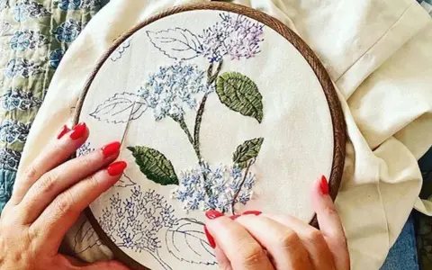 Manos con uñas rojas bordando una flor de hortensia en un bastidor. Se ve lo que se necesita para bordar: tela, hilo y aguja.
