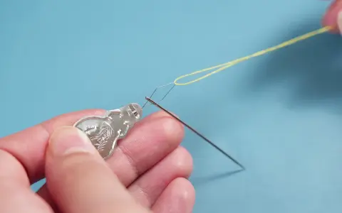 Manos usando un enhebrador de agujas para pasar hilo amarillo por el ojo de una aguja.