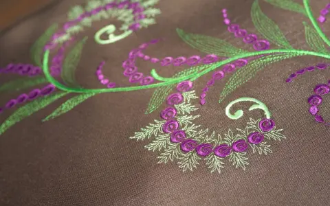 Detalles de bordado floral en tonos verdes y morados sobre tela marrón. Ideal para descargar letras para bordar a maquina y crear diseños únicos.