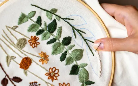 Manos creando bellas ideas para bordar: hojas verdes y flores naranjas en tela, con aguja e hilo listos para continuar.