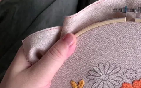 Mano sujetando un bastidor de bordar con lino para bordar. Se aprecian flores a medio bordar en tonos grises, naranjas y amarillos.