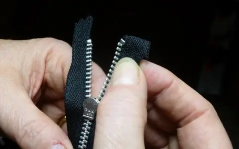 Manos sujetan una cremallera negra, alineando los dientes para coser cremallera a mano.