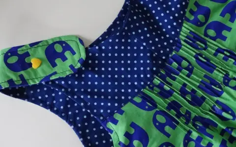 Detalle de un vestido infantil con estampado de elefantes azules sobre fondo verde. El cuello tiene un detalle de lunares azules y un botón amarillo en forma de corazón. Un adorable **patrón ranita bebé**.