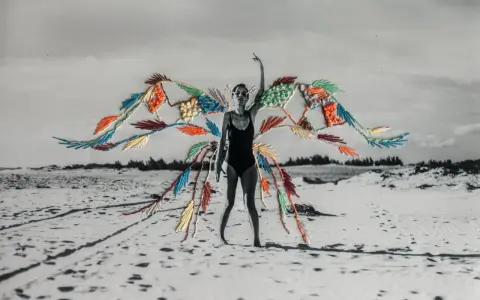 Mujer en traje de baño con alas de bordado de fotos coloridas sobre un paisaje de playa desértica.
