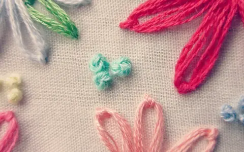 Detalle de bordado con flores de hilo. Se aprecian pétalos en rosa, rojo, azul y blanco, y un centro de nudo francés bordado en turquesa.