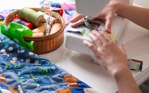 Manos cosiendo en máquina, con un costurero lleno de hilos y tijeras, y un quilt colorido.