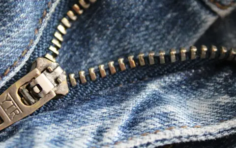 Primer plano de una cremallera atascada con tela de mezclilla azul. El tirador metálico muestra la marca "YTR".