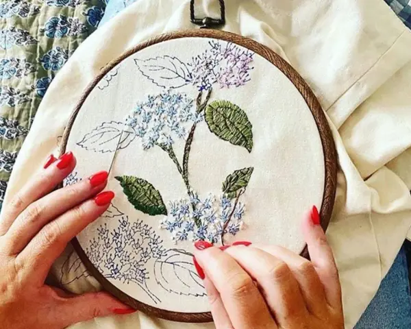 Manos con uñas rojas bordando una flor de hortensia en un bastidor. Se ve lo que se necesita para bordar: tela, hilo y aguja.