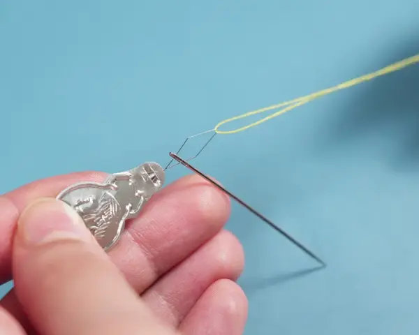 Manos usando un enhebrador de agujas para pasar hilo amarillo por el ojo de una aguja.