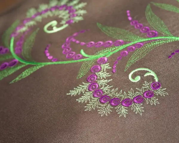 Detalles de bordado floral en tonos verdes y morados sobre tela marrón. Ideal para descargar letras para bordar a maquina y crear diseños únicos.