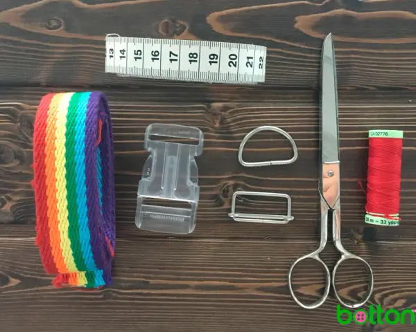 Herramientas para hacer un collar para perro: cinta métrica, cinta de arcoíris, hebilla, anillas, tijeras y hilo rojo.