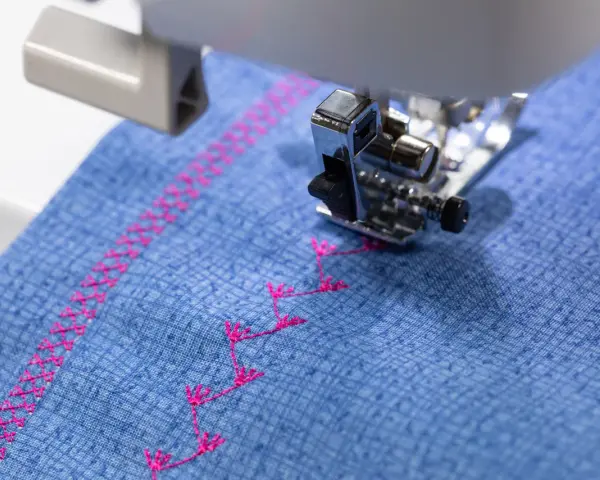 Máquina de coser bordando un patrón decorativo en **tejidos de algodón** azul con hilo rosa.