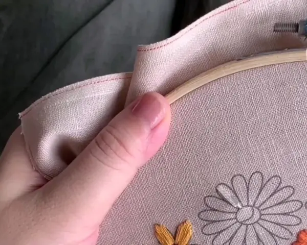 Mano sujetando un bastidor de bordar con lino para bordar. Se aprecian flores a medio bordar en tonos grises, naranjas y amarillos.