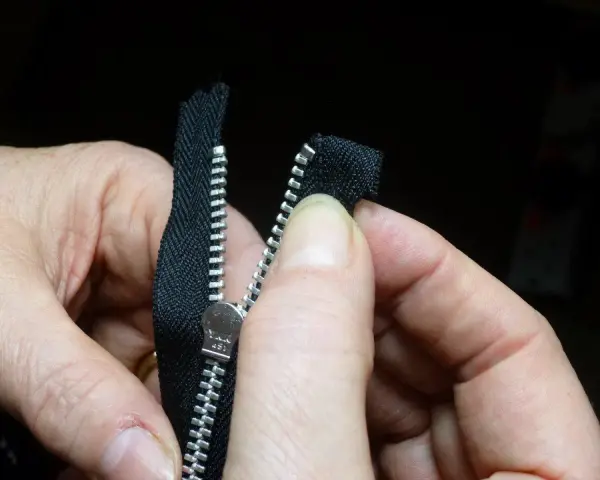 Manos sujetan una cremallera negra, alineando los dientes para coser cremallera a mano.