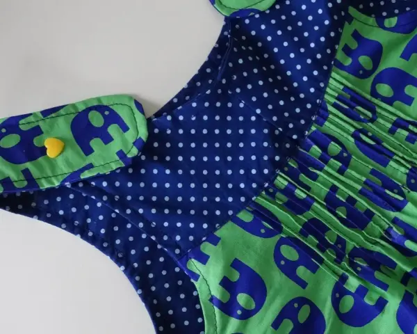 Detalle de un vestido infantil con estampado de elefantes azules sobre fondo verde. El cuello tiene un detalle de lunares azules y un botón amarillo en forma de corazón. Un adorable **patrón ranita bebé**.