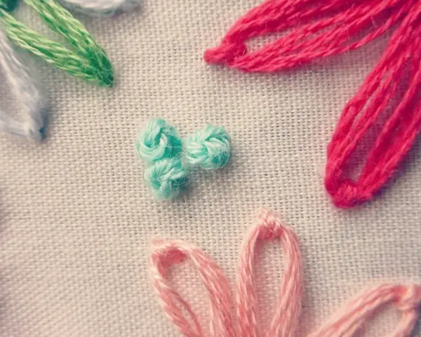 Detalle de bordado con flores de hilo. Se aprecian pétalos en rosa, rojo, azul y blanco, y un centro de nudo francés bordado en turquesa.