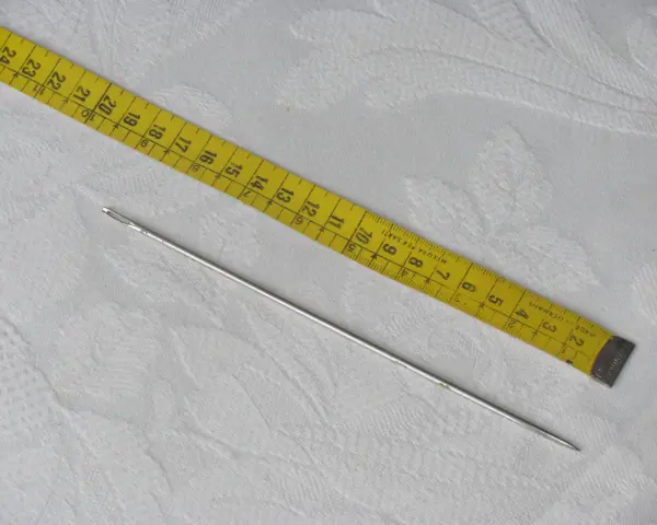 Una aguja de coser plateada y una cinta métrica amarilla, que muestra medidas en centímetros. La aguja, que es como un punzón fino, está lista para usarse.