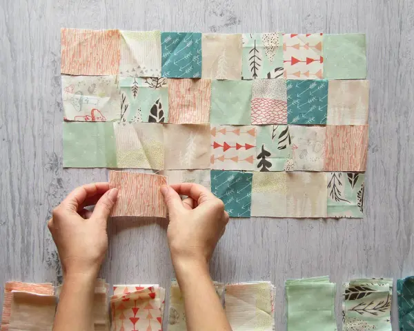 Manos uniendo retazos de tela con diseños de árboles y flechas, creando un patrón de patchwork.