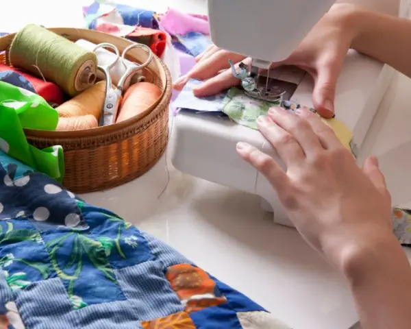 Manos cosiendo en máquina, con un costurero lleno de hilos y tijeras, y un quilt colorido.