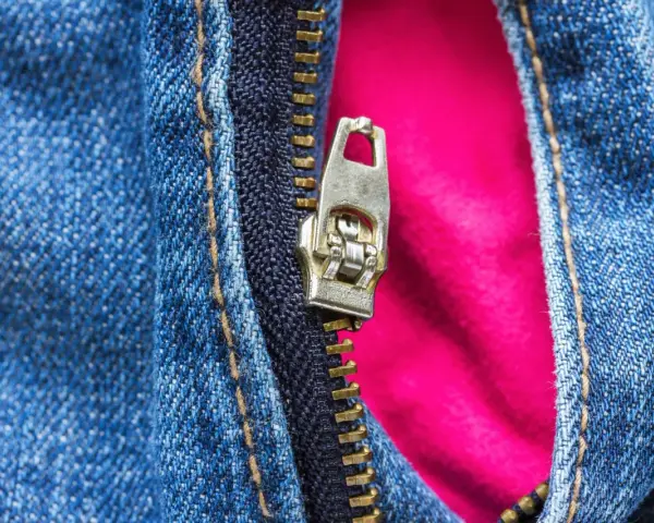 Detalle de cremallera metálica en jeans azules, con un forro rosa vibrante que se asoma. La cremallera salida de un lado revela el contraste de texturas.