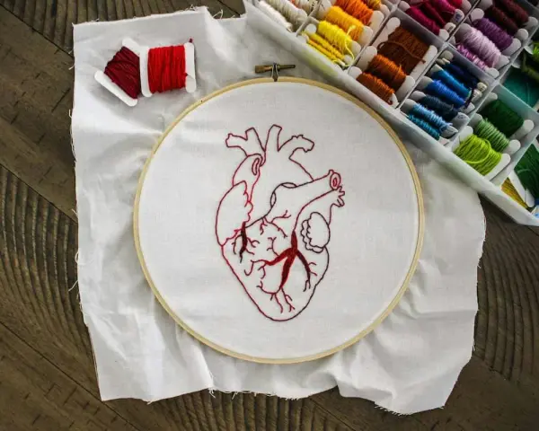 Un corazón bordado en tela blanca, listo para aprender a bordar. Hilos rojos y una caja de hilos de colores completan la escena.